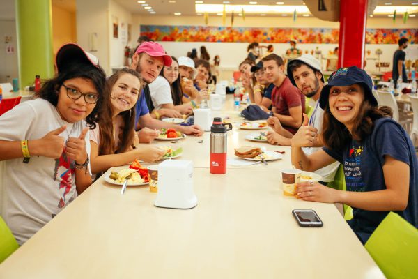 קבוצת חניכים ומדריכים יושבים לאורך שולחן ארוך בחדר האוכל
