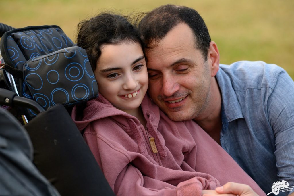 ילדה המתניידת בכסא גלגלים מחייכת ומאחוריה אבא שלה עם עיניים עצומות מחבק אותה